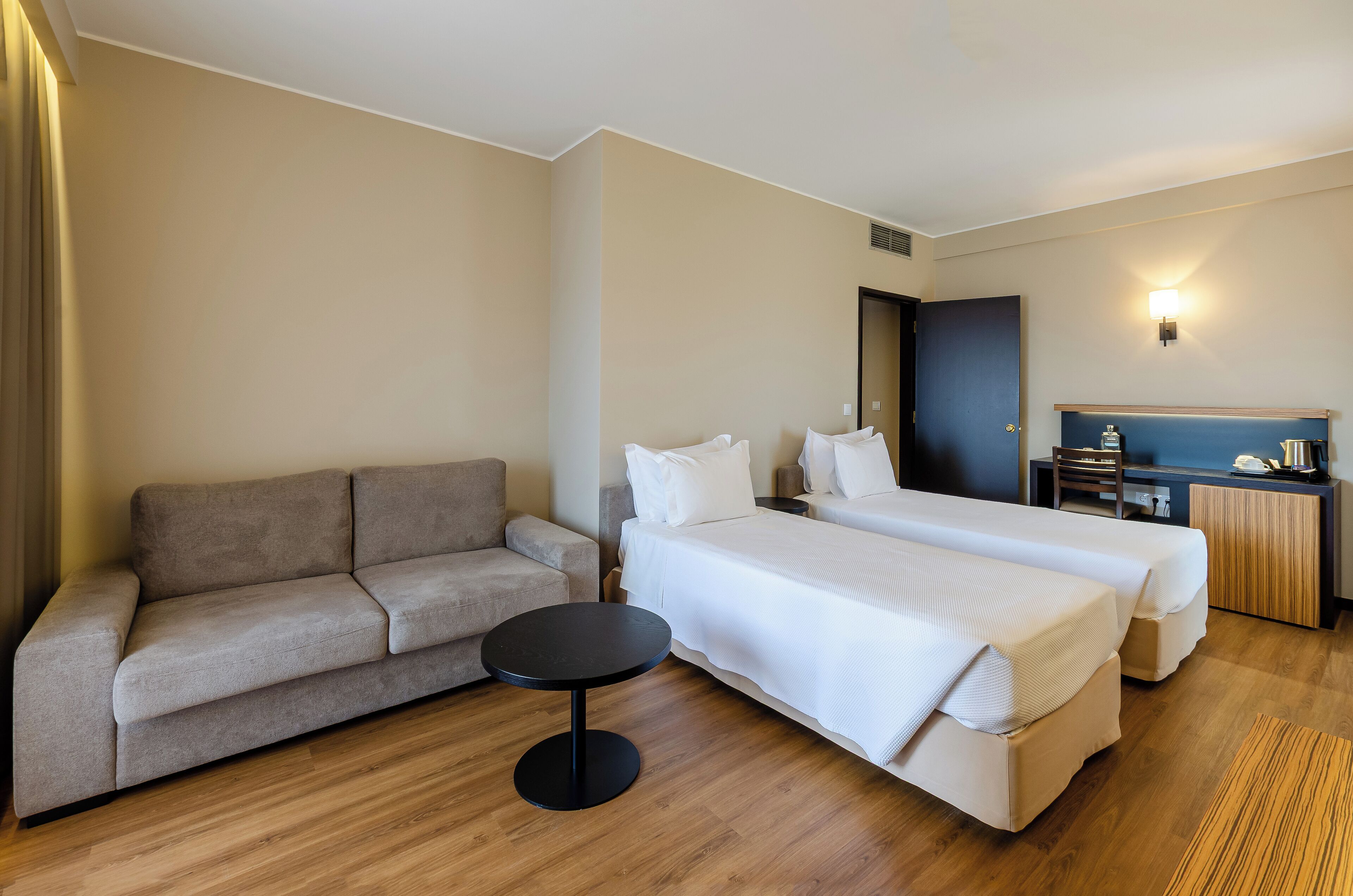 A hotel room with a sofa, two single beds, a small round table, and a minibar.