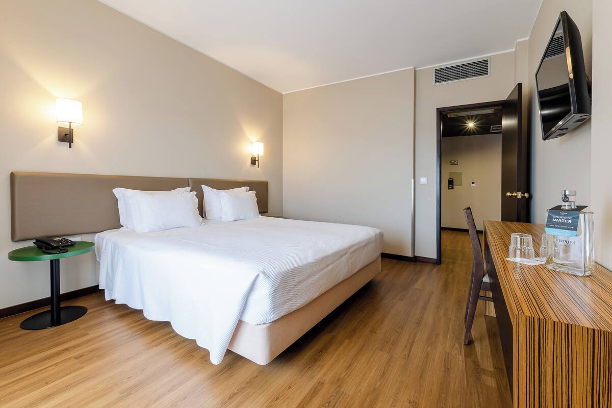 Une chambre d’hôtel avec un lit, une table de chevet, une lampe et un bureau en bois sur lequel se trouve une bouteille d’eau.