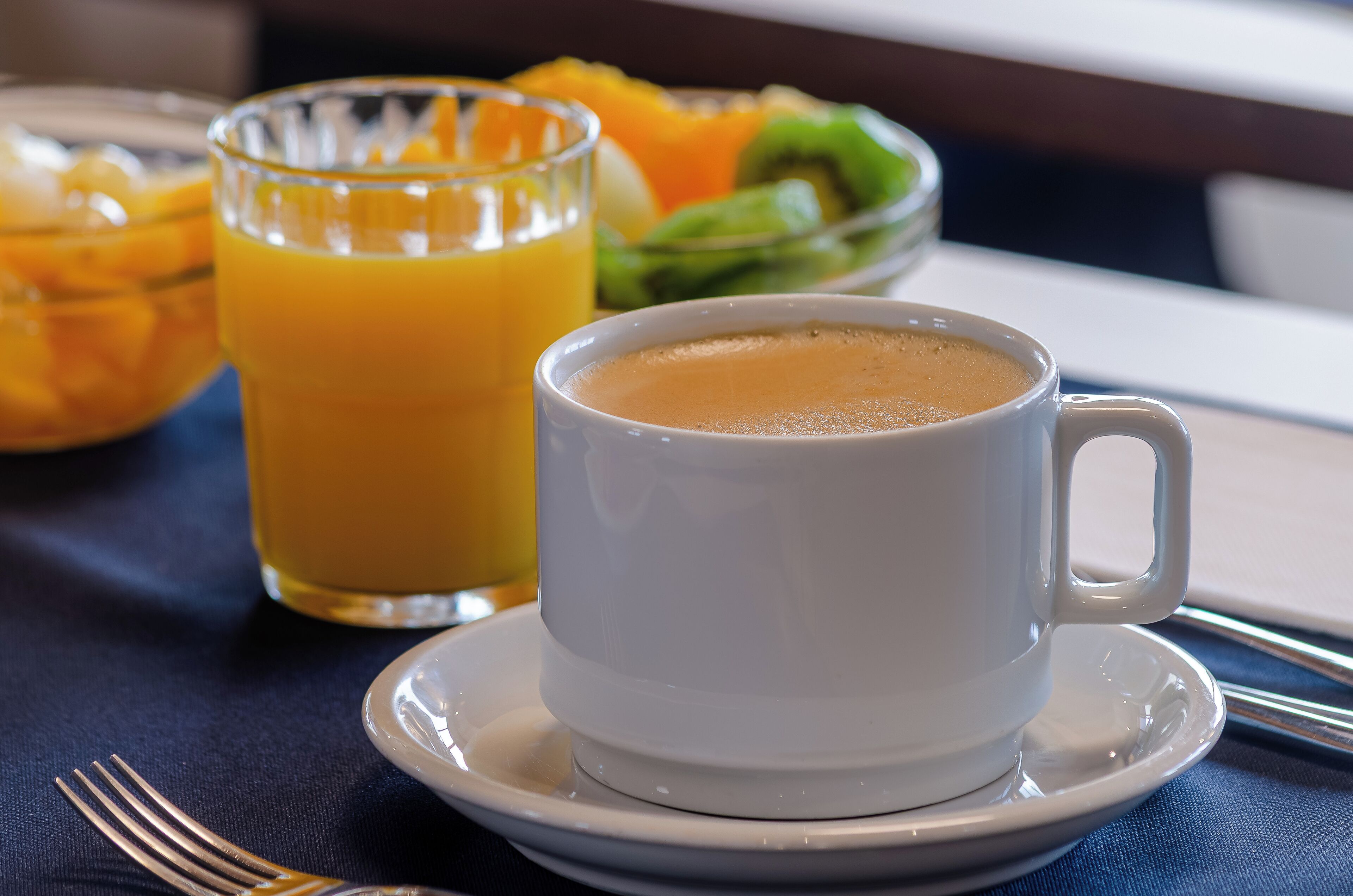 A white cup of coffee on a saucer, a glass of orange juice, and a bowl of fruit on a table.