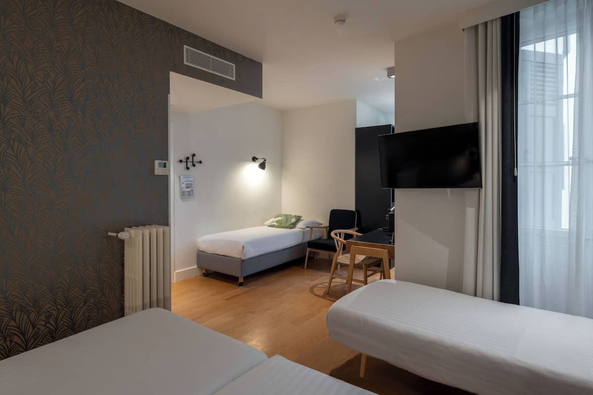 A hotel room with two single beds, a desk with a chair, a TV mounted on the wall, and a window with curtains.