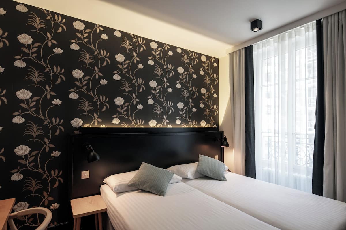 A hotel room with a black floral wallpaper, a black headboard, two beds with white bedding and grey pillows, a wooden nightstand, and a window with sheer curtains.