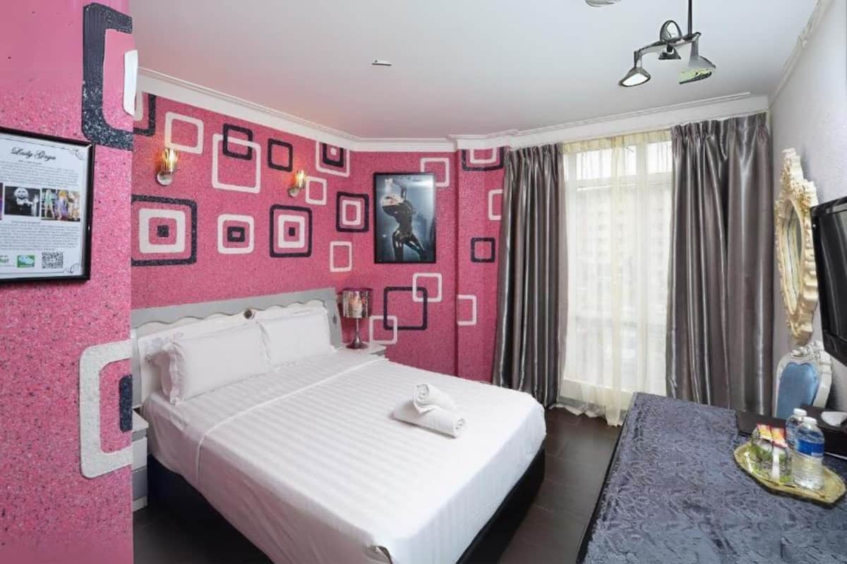 Quarto de hotel com uma cama grande, uma escrivaninha e uma cadeira. Há uma janela com cortinas, um quadro na parede e uma fotografia emoldurada na parede.