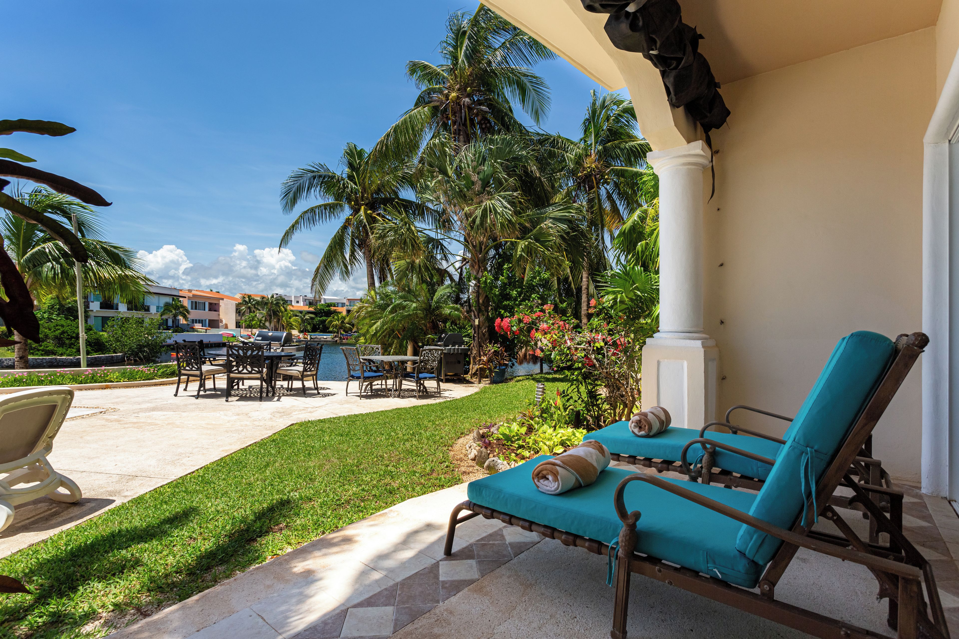 An outdoor patio with lounge chairs, a dining table, and chairs, surrounded by palm trees and a pool.