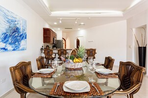 Un comedor con una mesa de vidrio rodeada de sillas de madera, un centro de mesa con frutas y una botella, y una cocina moderna al fondo.
