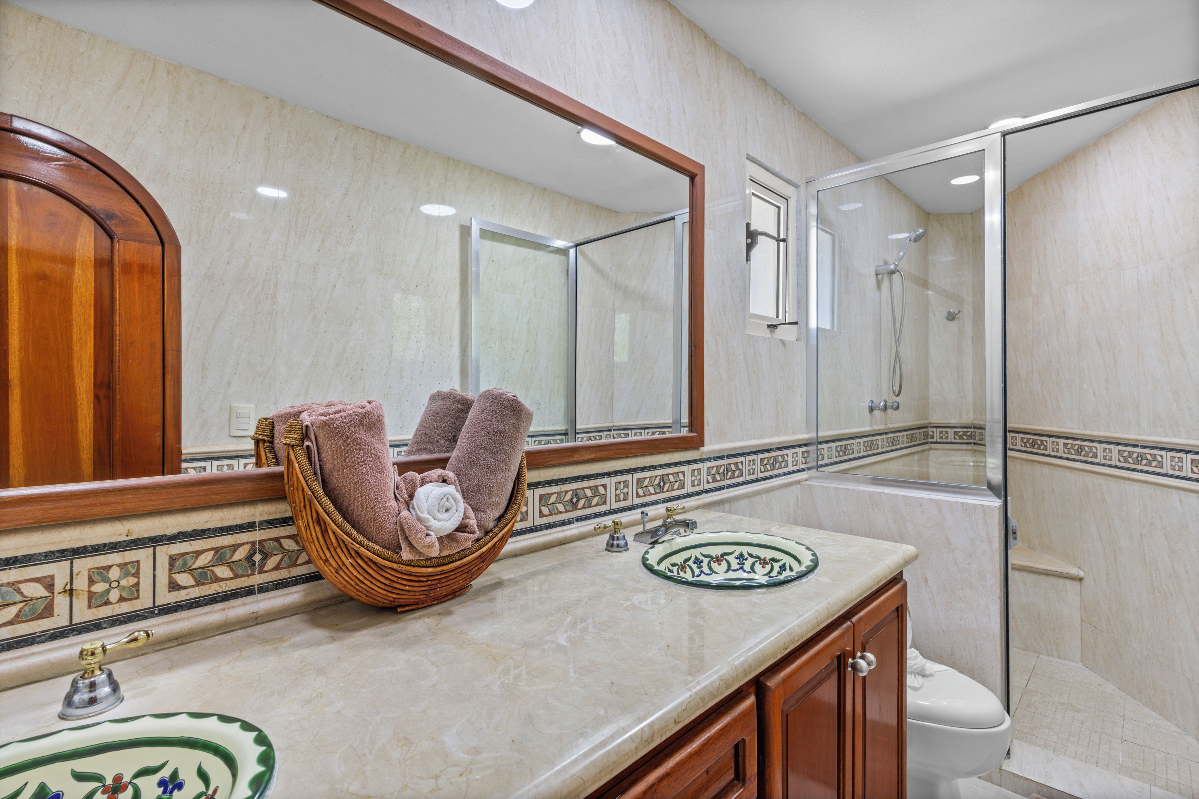 Une salle de bain dotée d’un comptoir en marbre, d’un meuble-lavabo en bois, d’une cabine de douche en verre et d’un miroir décoratif.