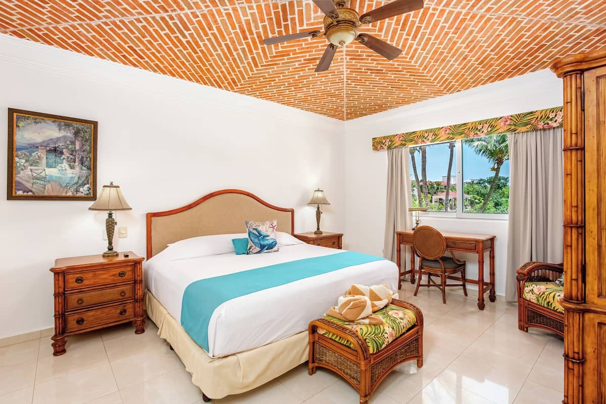 A hotel room with a bed, two bedside tables, a desk with a chair, a window with curtains, and a ceiling fan.