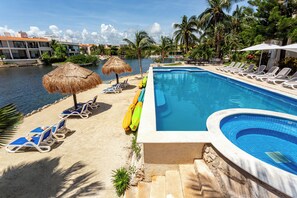 A swimming pool with lounge chairs and umbrellas, surrounded by palm trees and a body of water. - Aventuras Club Lagoon (Puerto Aventuras)