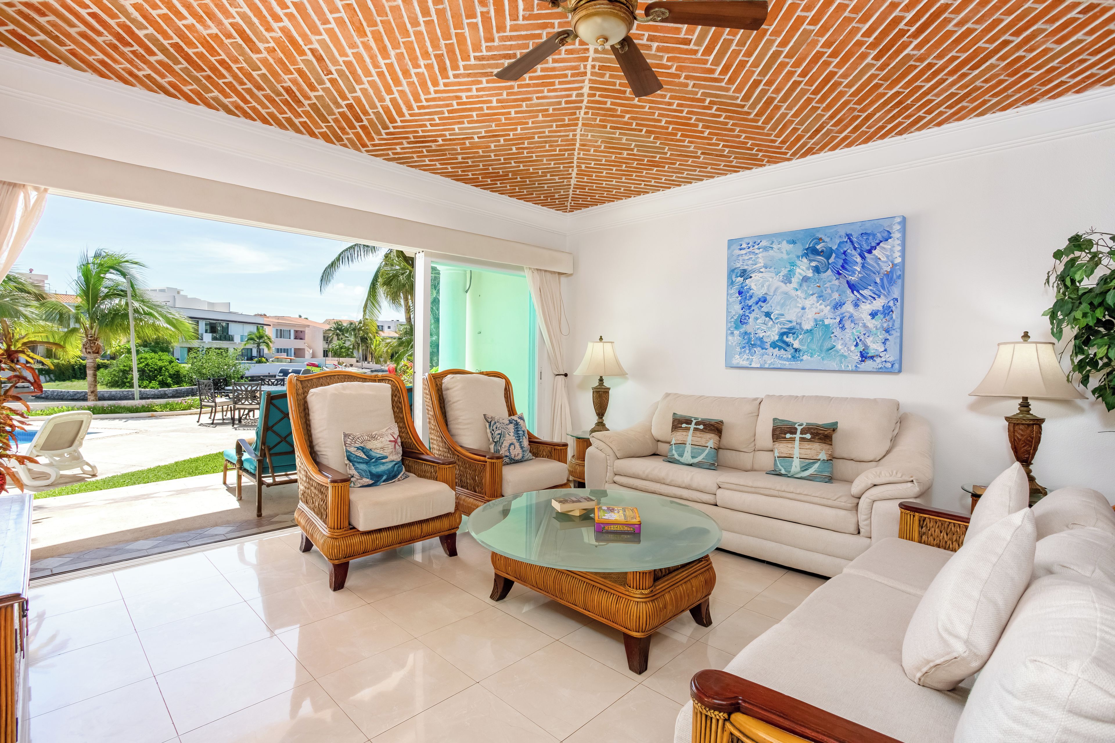 A spacious living area with a ceiling fan, a glass coffee table, wicker furniture, a sofa with decorative pillows, and a view of a pool and palm trees outside.