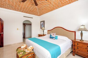 Un dormitorio con techo de ladrillo, una cama con colcha azul, mesitas de noche de madera y una silla de mimbre.