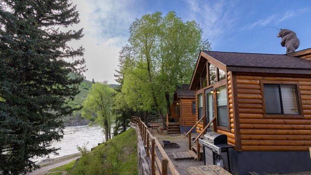 A log cabin with a deck overlooking a river, surrounded by trees. - Snake River Cabin Village (Jackson)