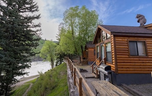 A log cabin with a deck overlooking a river, surrounded by trees. - Snake River Cabin Village (Jackson)