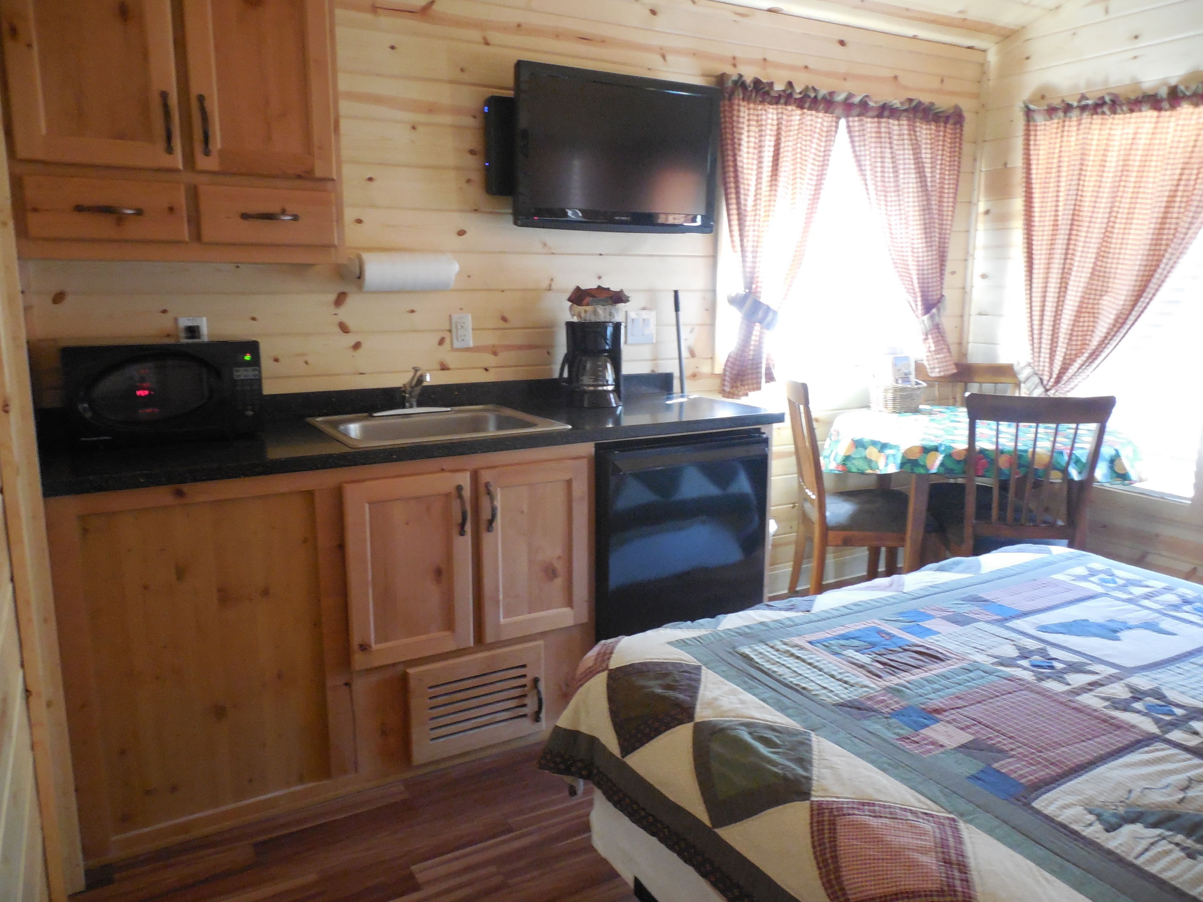 A compact cabin-style room with wooden walls, a kitchenette, a bed with a quilt, and a television mounted above the counter.