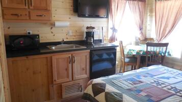 A compact cabin-style room with wooden walls, a kitchenette, a bed with a quilt, and a television mounted above the counter.