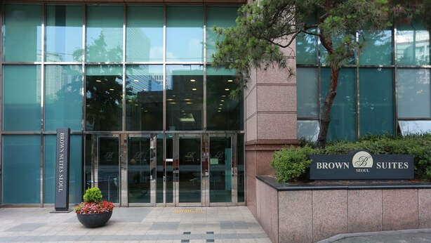 A hotel entrance with a sign reading "Brown Suites Seoul," a glass door entrance, and a planter with flowers. - Brown Suites Seoul (Seoul)