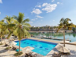 Una piscina con sillas de descanso y sombrillas, un puerto deportivo con barcos, y palmeras.