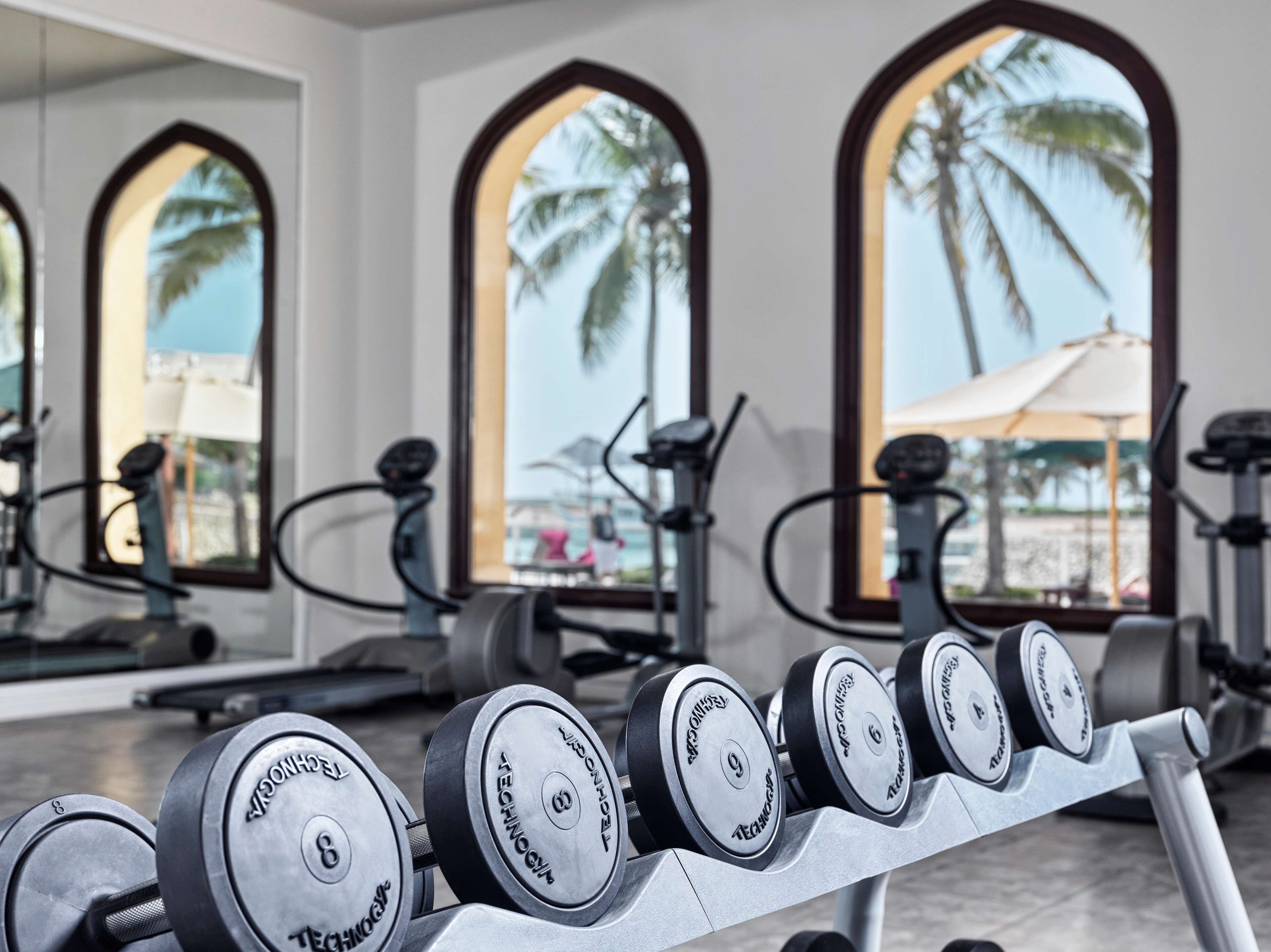 A gym with exercise equipment and large windows with views of palm trees and a beach.