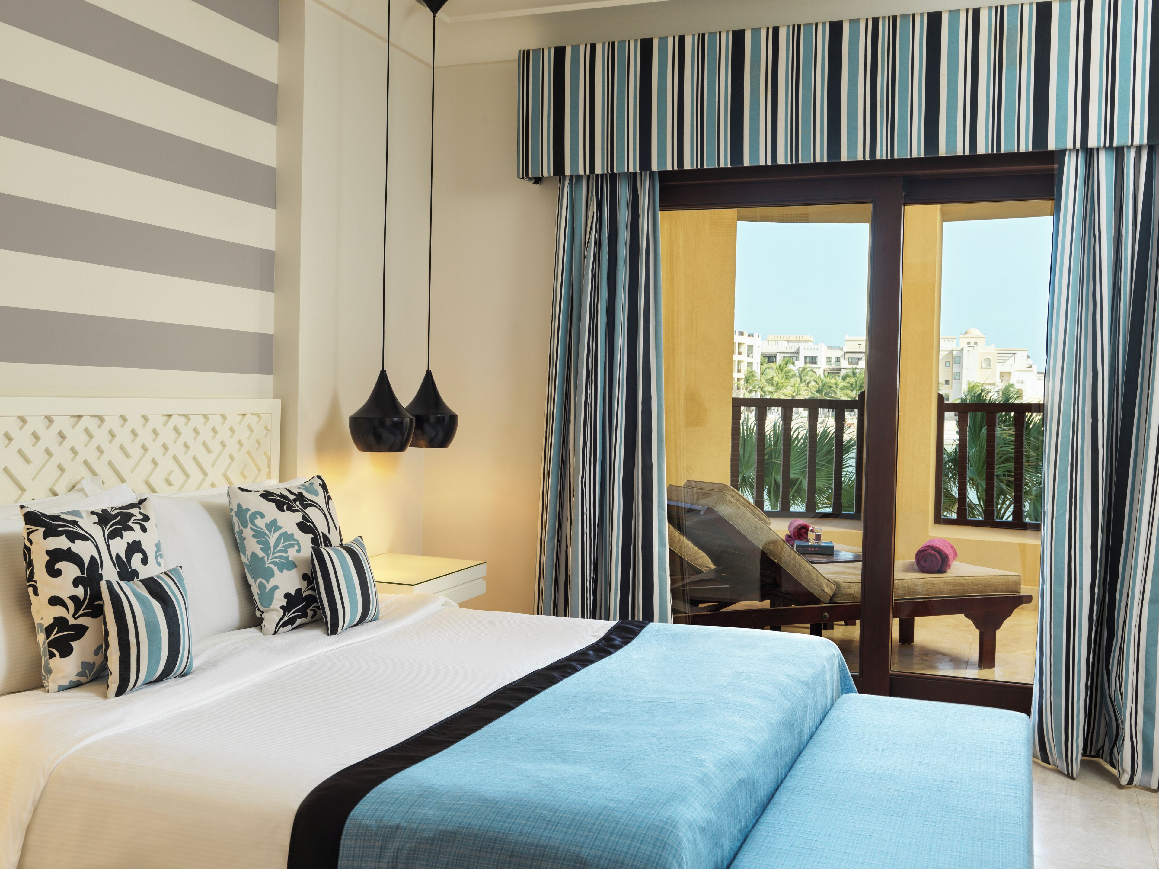 A hotel room with a large bed, a nightstand, two pendant lights, a view of the balcony with a chair and a table, and a striped curtain.