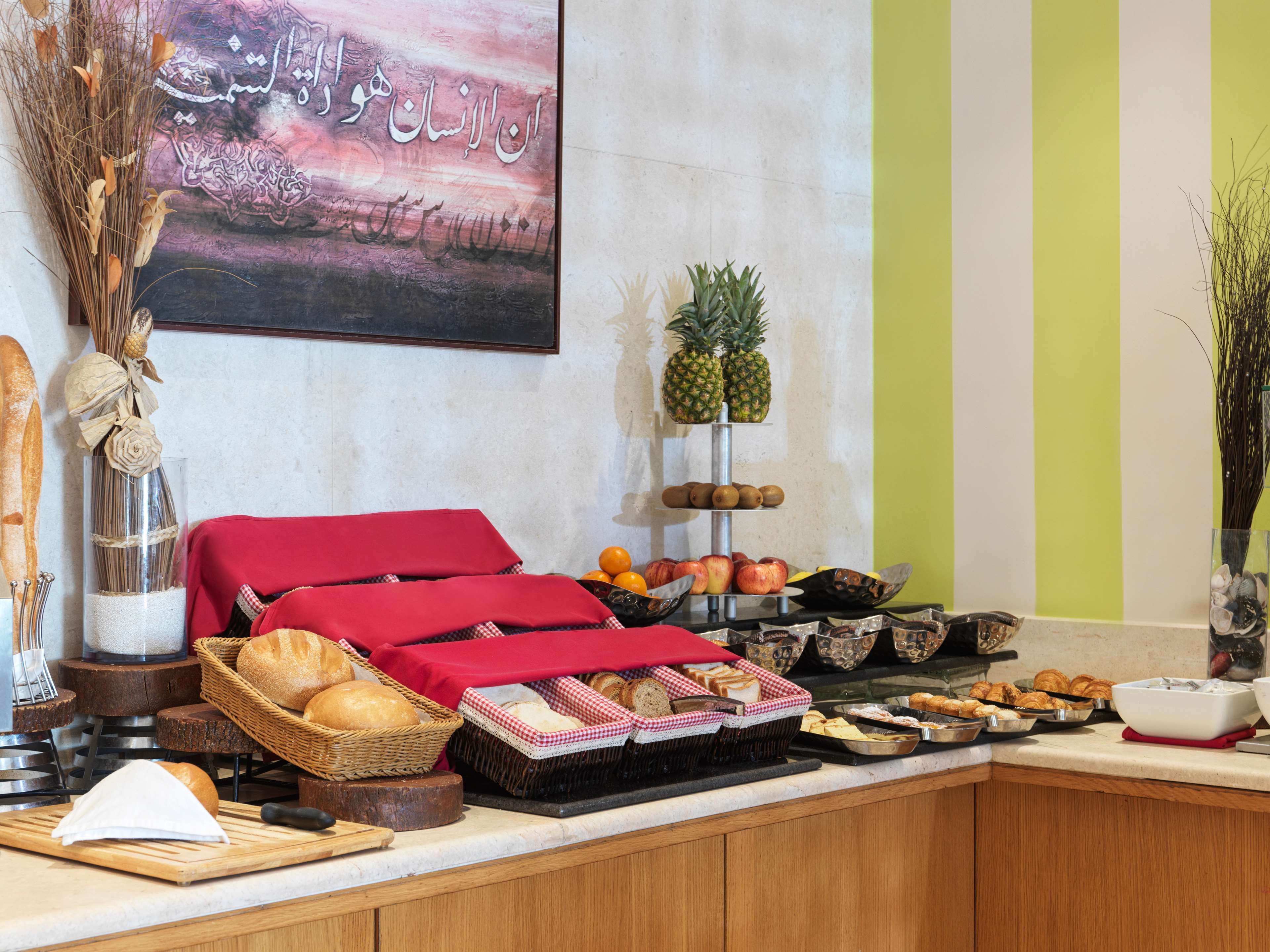 A breakfast buffet with bread, pastries, fruits, and a decorative wall with Arabic calligraphy.