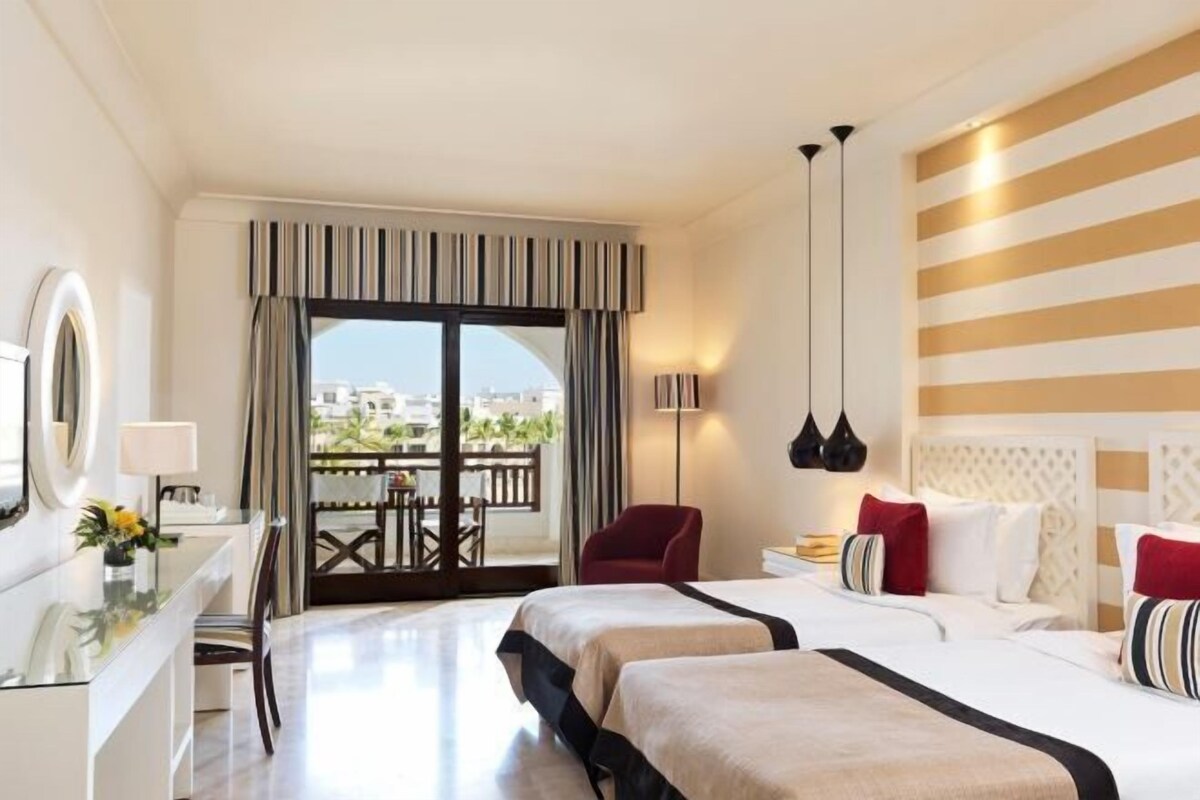 A hotel room with a bed, a desk, a chair, a lamp, and a view of the outdoors.