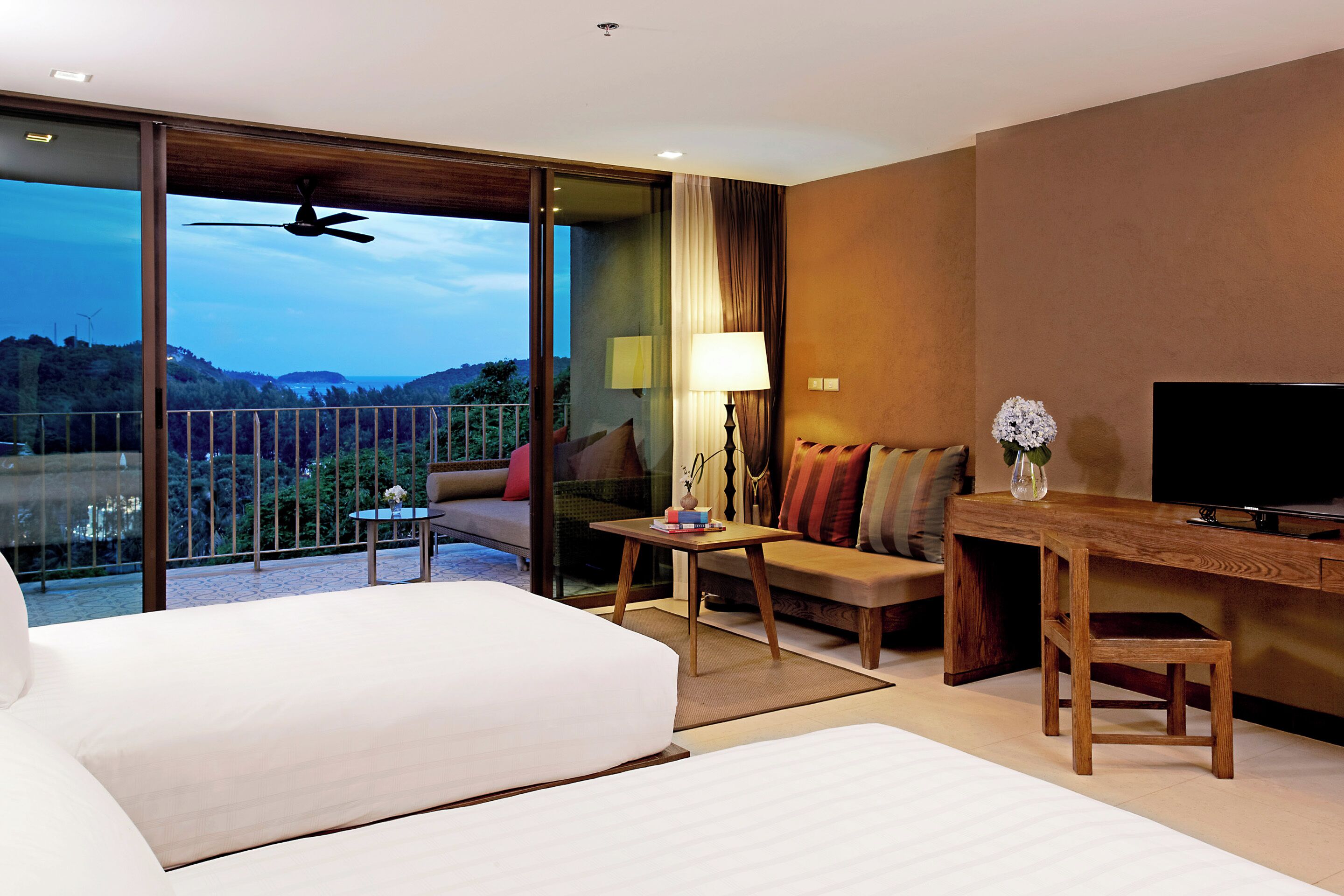 A hotel room with a large bed, a desk, a chair, a sofa, a TV, and a balcony with a view of the hills.
