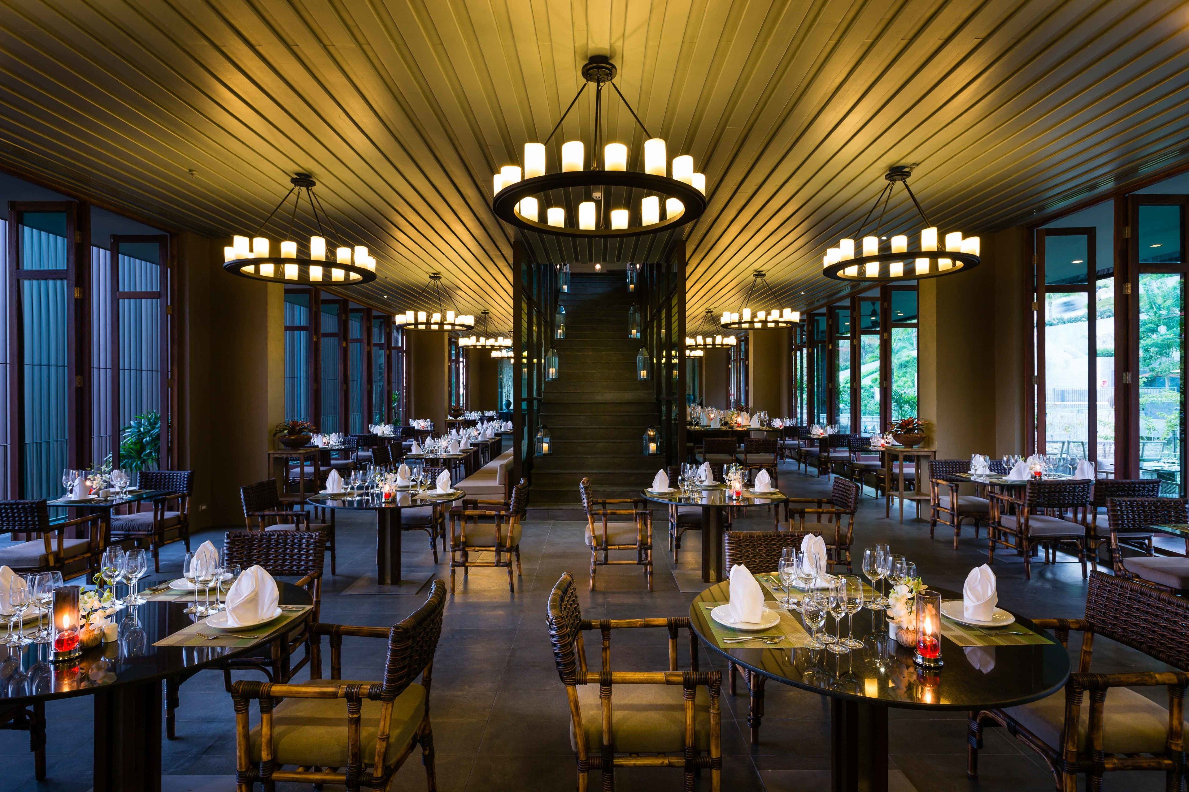 A spacious dining area with multiple tables set for guests, featuring a central staircase and large windows providing natural light.