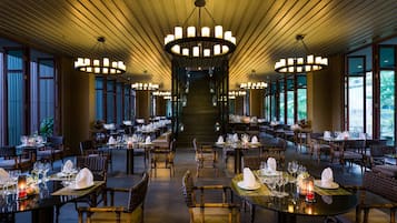 A spacious dining area with multiple tables set for guests, featuring a central staircase and large windows providing natural light.