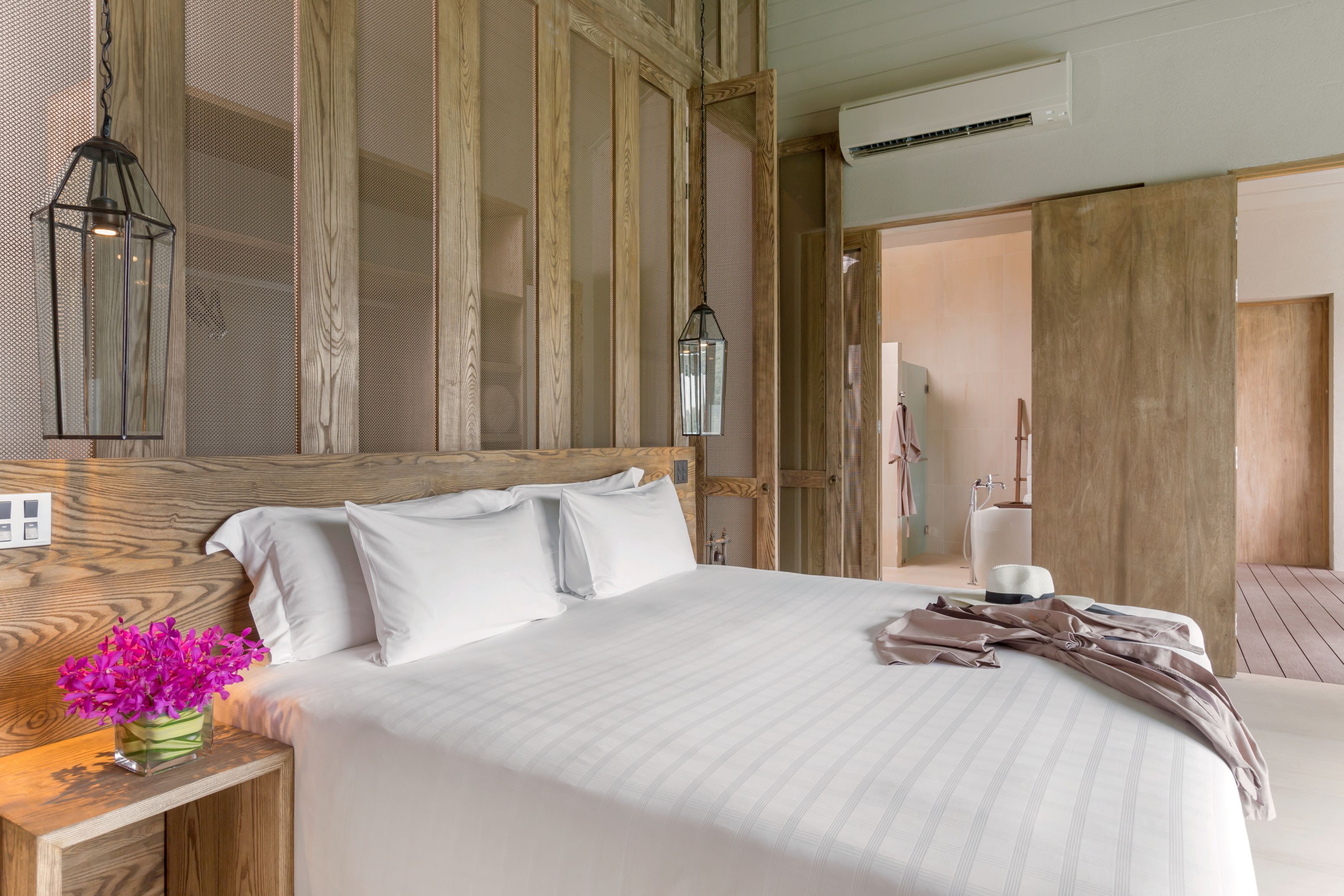 A hotel room with a large bed, wooden headboard, two bedside lamps, a nightstand with a vase of flowers, and a wooden door leading to another room.