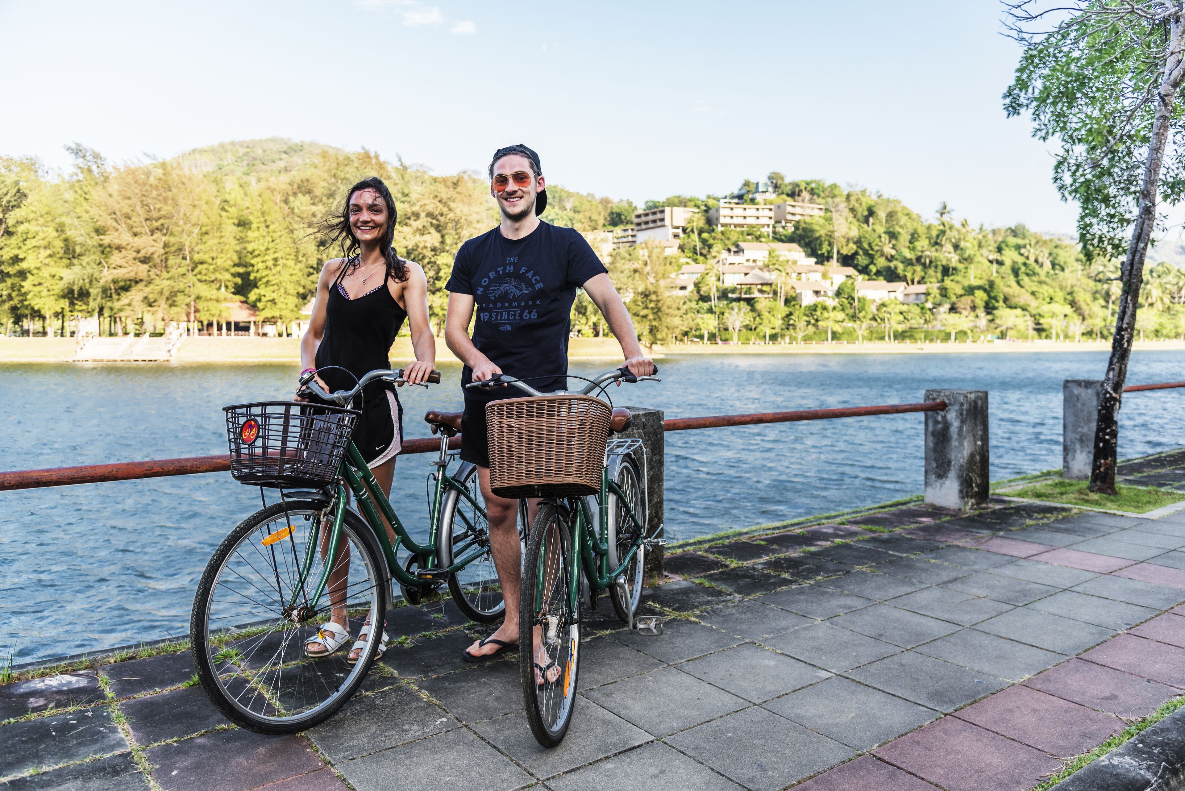 Two people standing by bicycles on a waterfront promenade with a view of a lake and buildings in the background.