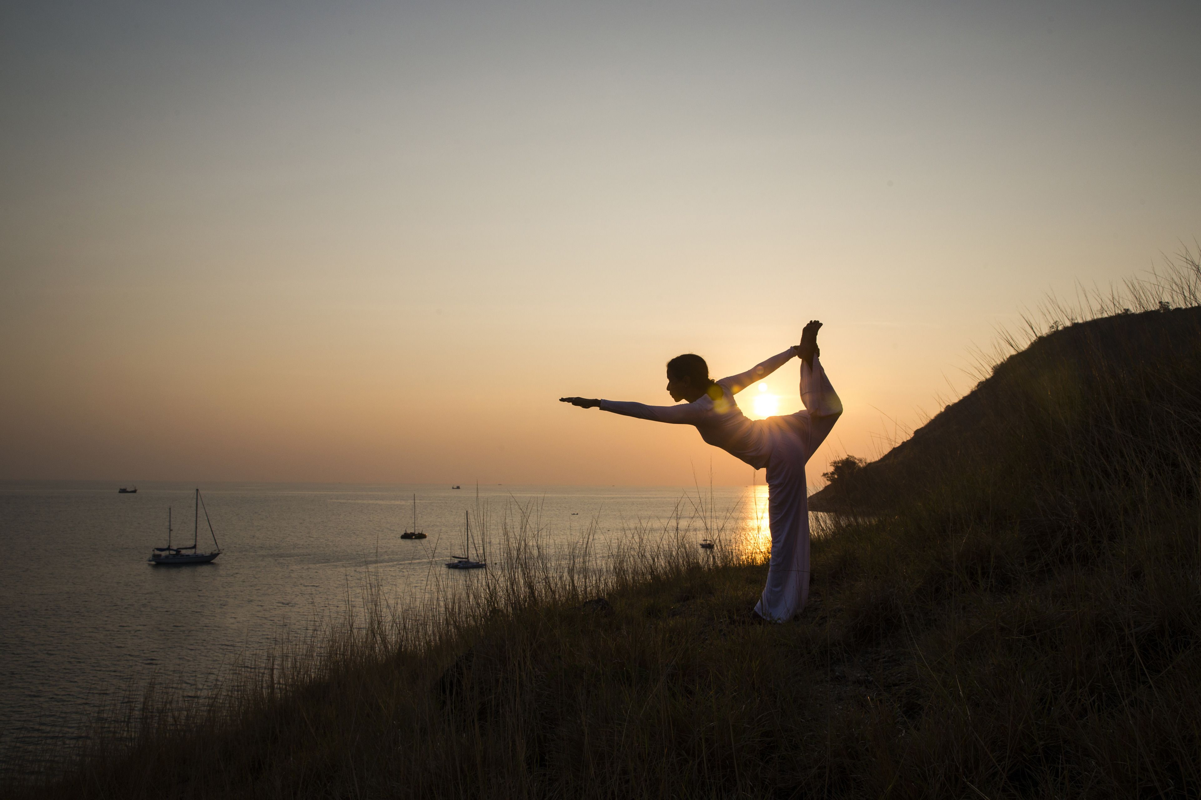 Una persona realizando una postura de yoga en una colina cubierta de hierba, con vista al mar tranquilo y barcos.