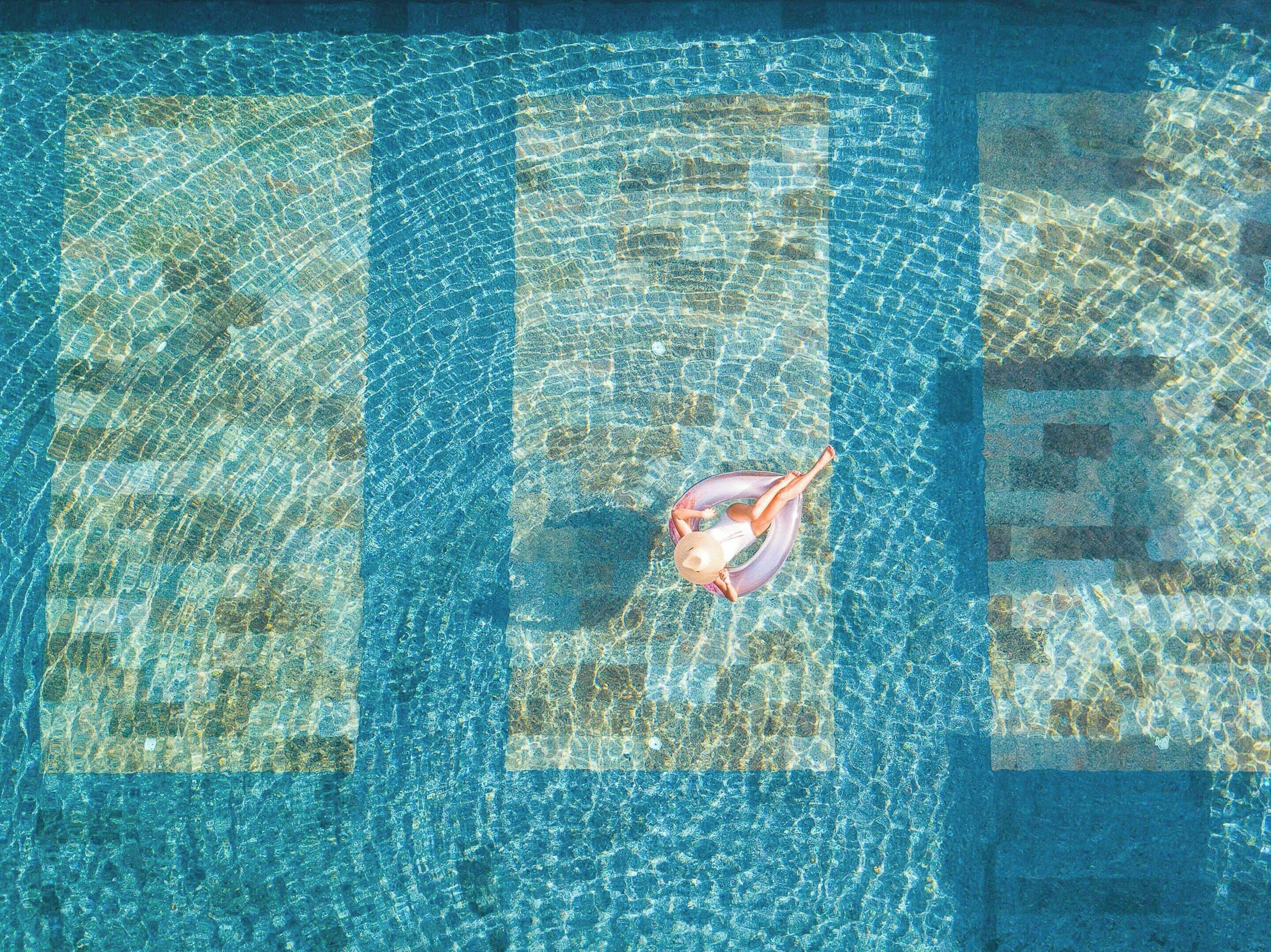 Eine Luftaufnahme eines Schwimmbeckens mit klarem Wasser, einer Person, die auf einem ringförmigen Schwimmdieling treibt, und einer gefliesten Poolterrasse darum herum.