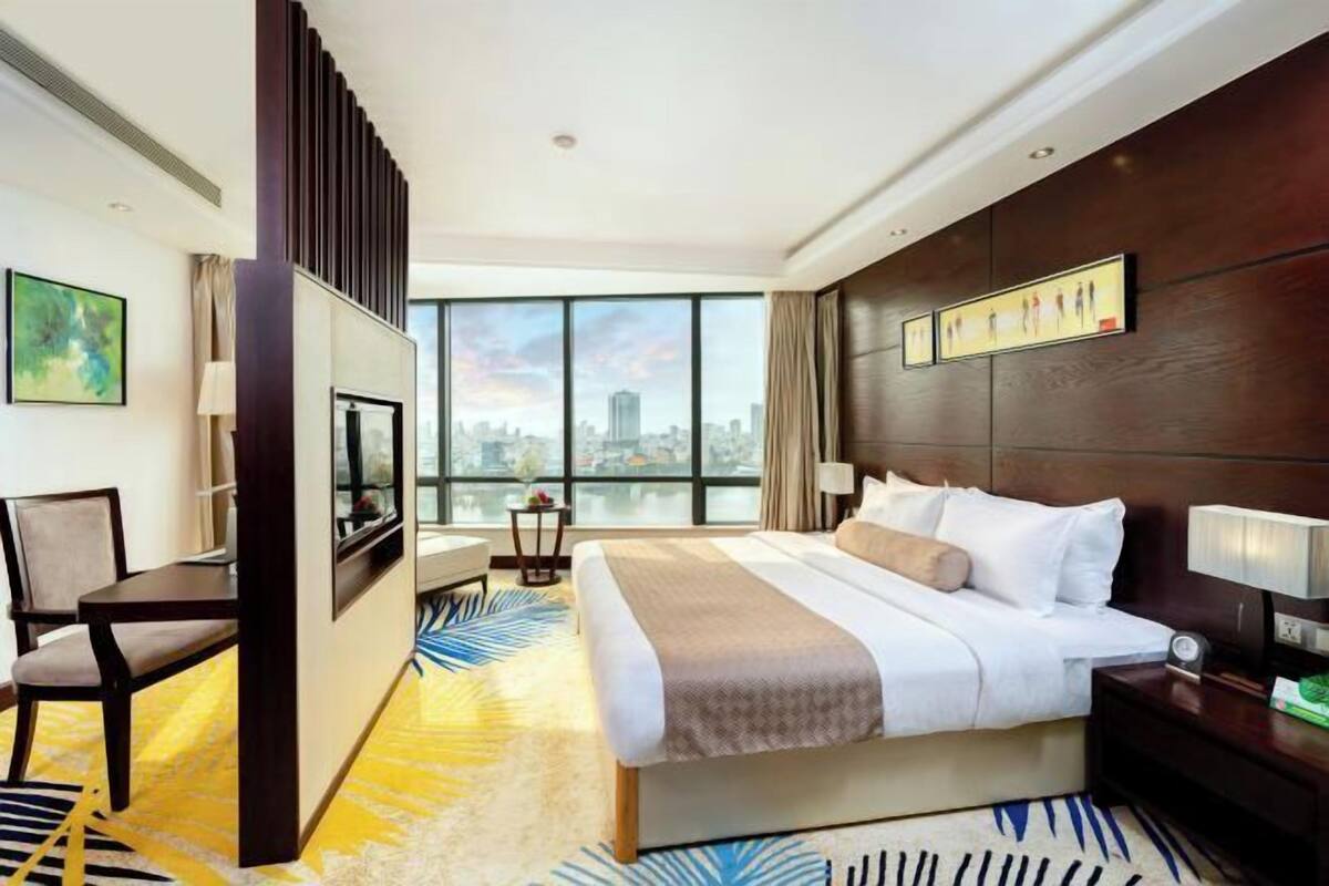 A hotel room with a bed, a TV, a desk, a chair, a window with a city view, and a large window.