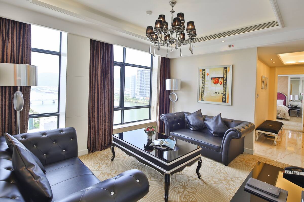 A modern hotel room with a large window, a chandelier, a sofa, a coffee table, and a painting on the wall.
