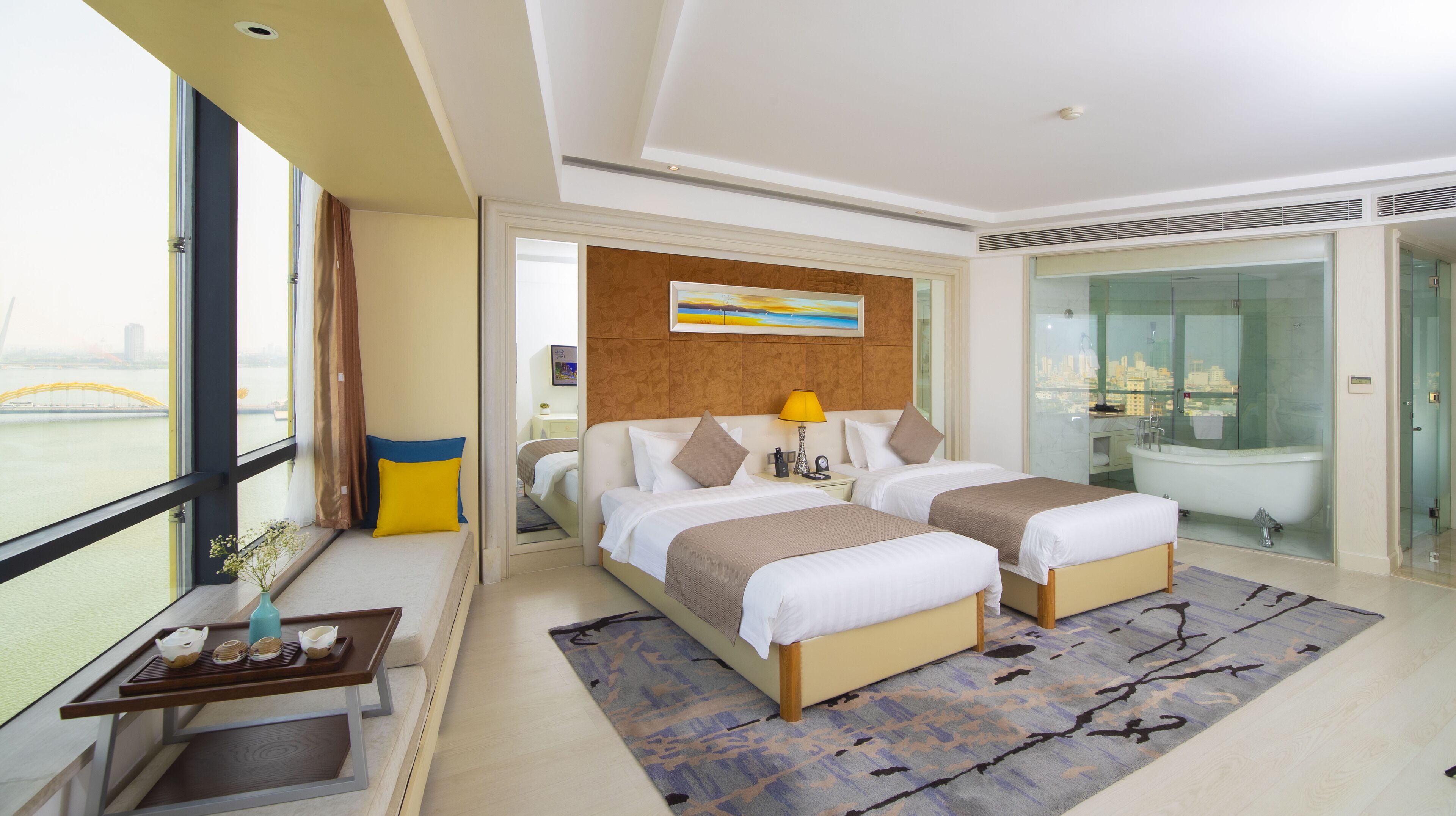 Una habitación de hotel moderna con dos camas, un ventanal amplio con vistas a la ciudad, un baño con ducha de vidrio y un área de comedor con una mesa y sillas.