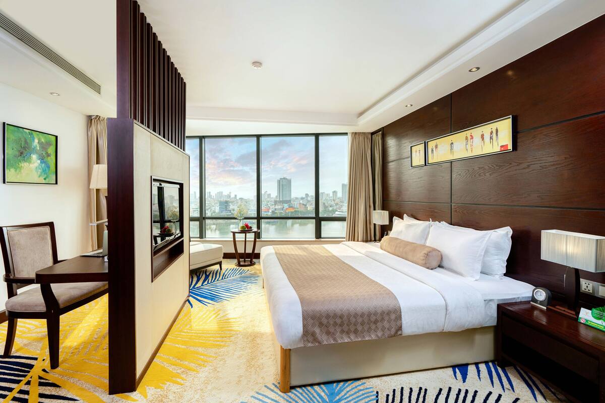 A hotel room with a large bed, a desk, a chair, a TV, and a view of the cityscape.