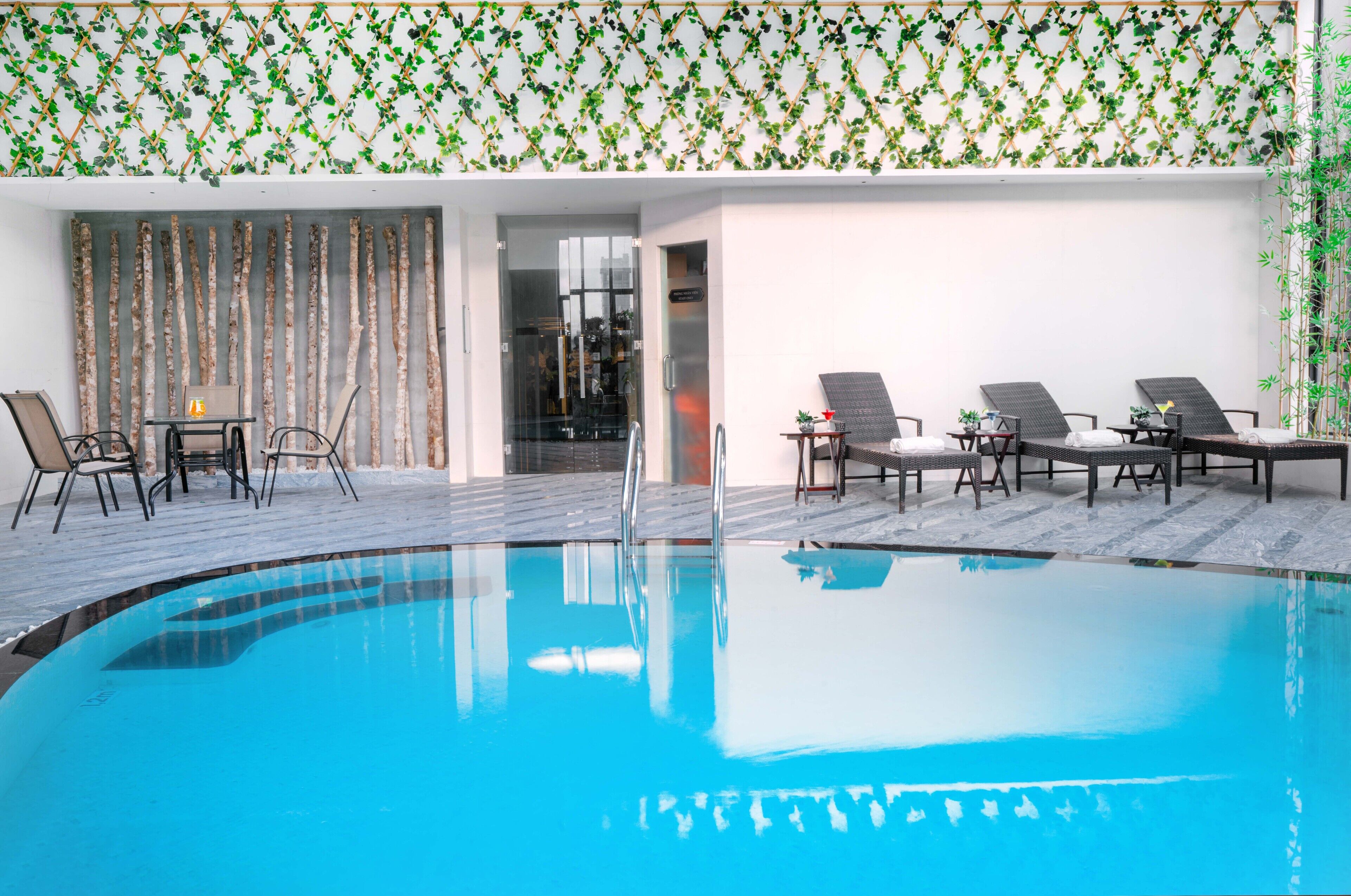 Un espace piscine doté de chaises longues et d’une table, entouré d’une terrasse en bois et d’un mur orné d’une plante grimpante.