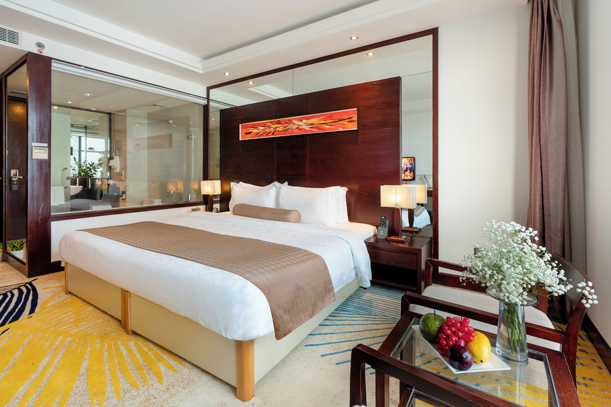 A hotel room with a large bed, bedside tables, a mirror, and a small table with fruit and a vase.