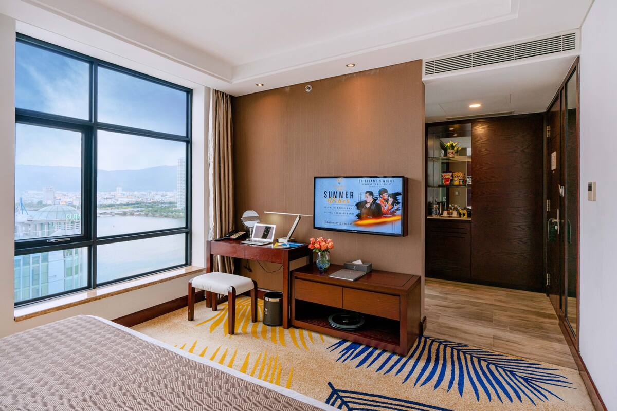 A hotel room with a large window, a desk with a laptop, a television, and a bed.
