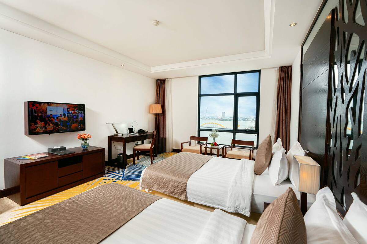 A hotel room with two beds, a desk, a TV, and a large window with a city view.
