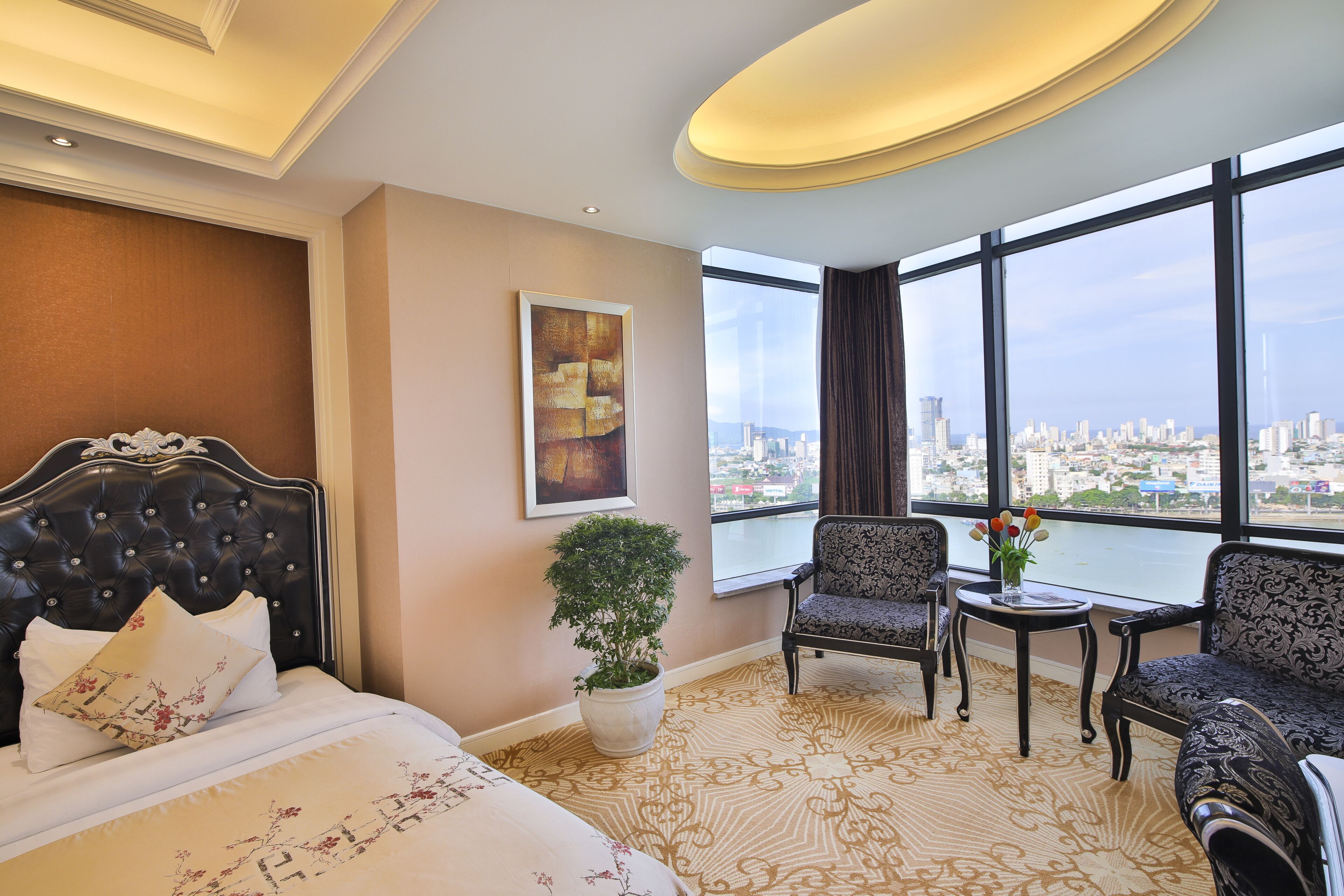A hotel room with a large bed, two armchairs, a small table with a vase, a potted plant, and a painting. The room has large windows with a city view.