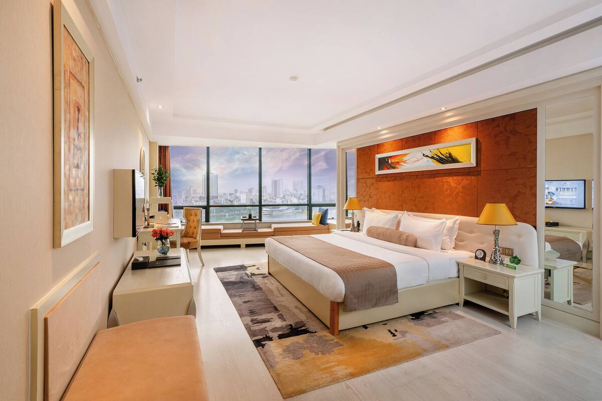 A modern hotel room with a large bed, a desk, a chair, and a view of the cityscape through large windows.