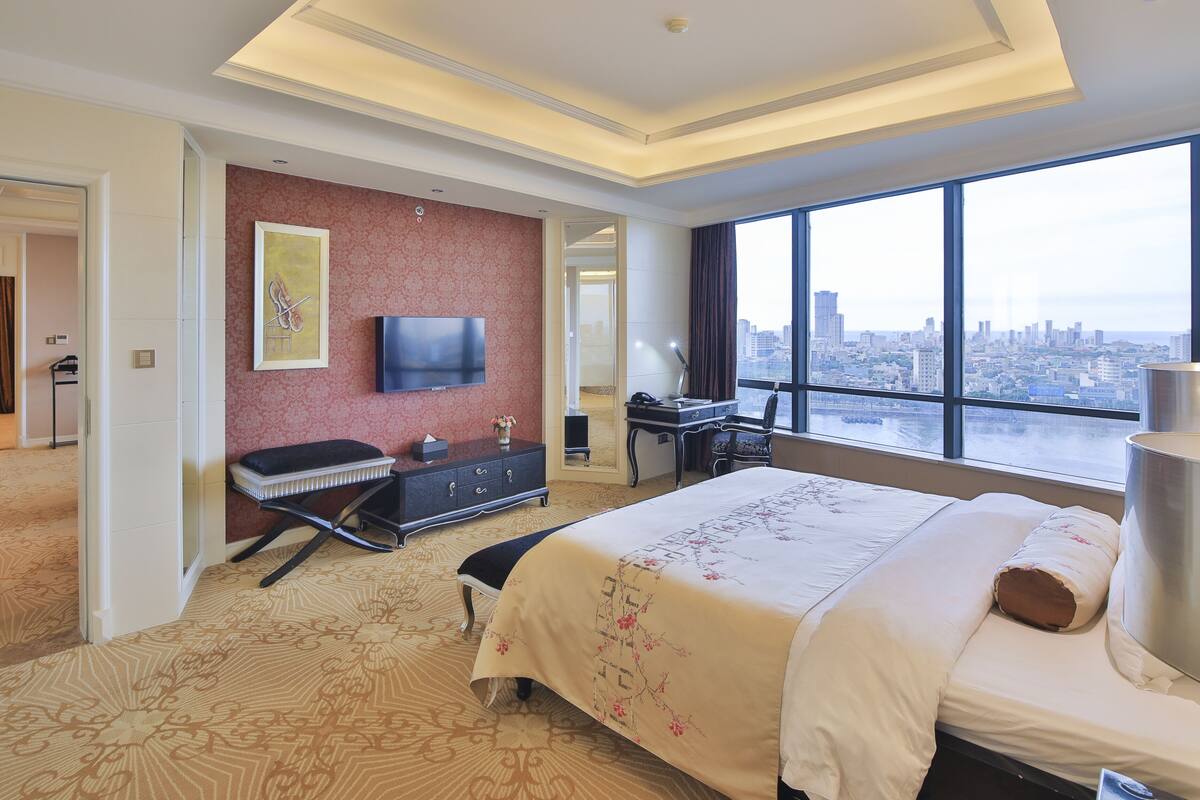 A hotel room with a large bed, a desk with a chair, a TV mounted on the wall, and a view of a cityscape through a large window.