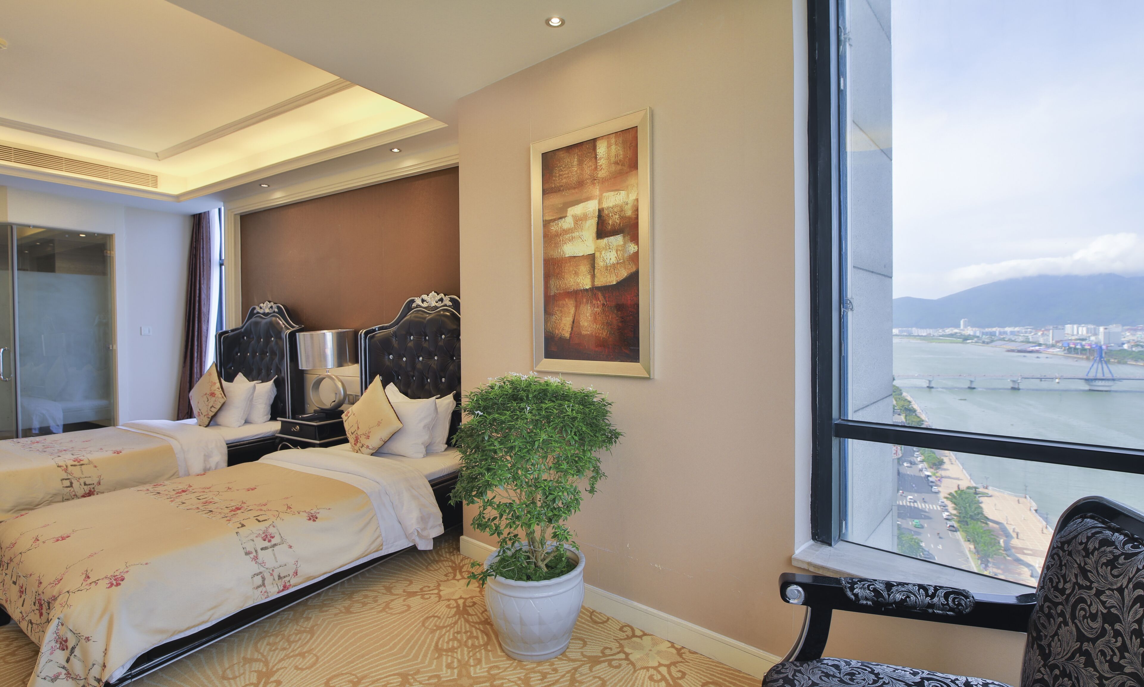 Habitación de hotel con dos camas, un ventanal grande con vista a la ciudad, una planta en maceta y un cuadro abstracto enmarcado.