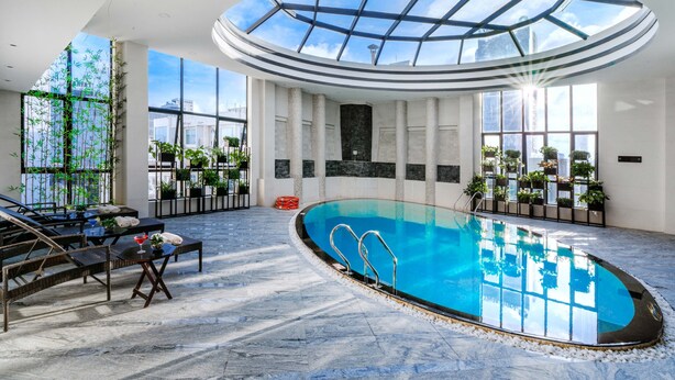 An indoor swimming pool with a glass ceiling, surrounded by seating areas and potted plants. - Brilliant Hotel (Da Nang)