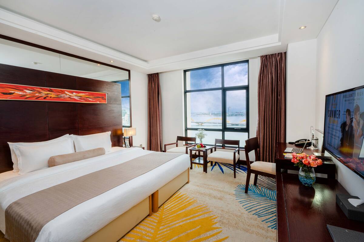 A hotel room with a large bed, a desk with a laptop, a TV, and a view of the city through the window.