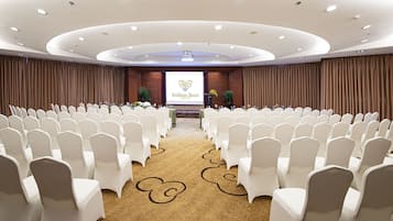 A conference room with rows of white chairs, a carpet with a floral pattern, and a screen displaying the hotel name and logo.
