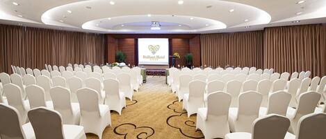 Un salón de conferencias con filas de sillas blancas, una alfombra con estampado floral y una pantalla que muestra el nombre y el logotipo del hotel.