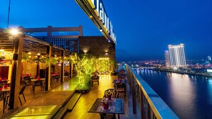 A waterfront restaurant with outdoor seating, a view of the city skyline, and a prominent illuminated sign. - Brilliant Hotel (Da Nang)