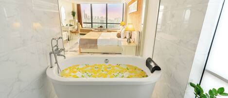 A modern bathroom with a large bathtub, a city view, and a cozy sitting area with a bed and lamps.