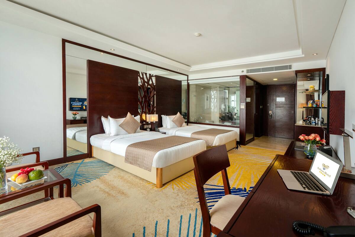 A hotel room with two beds, a desk with a laptop, a chair, a small table with fruit, and a mirror.