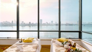 A hotel room with two beds, a large window with a city view, and a small table with a bottle and a towel. - Brilliant Hotel (Da Nang)